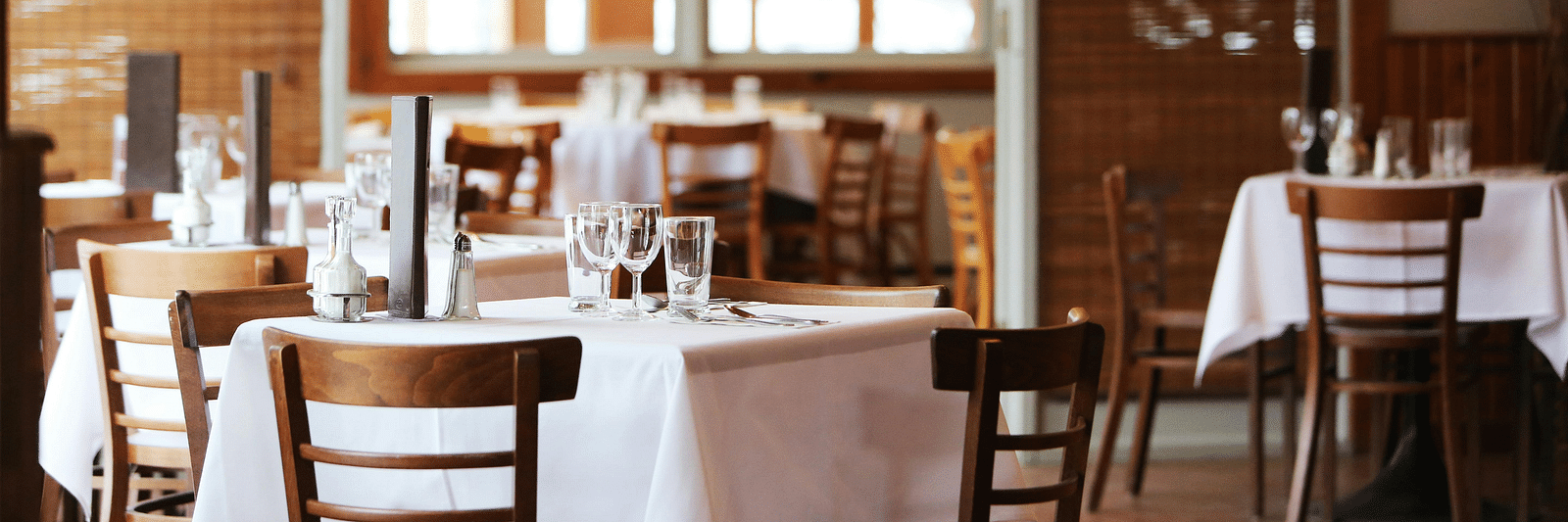 Empty tables set for dining in a restaurant with large windows.